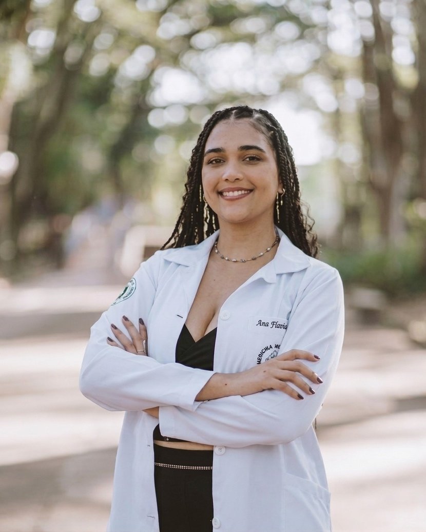 Ana Flávia, de jaleco branco e braços cruzados, sorrindo em uma alameda arborizada